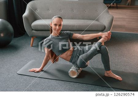 Sitting on the foam roller and smiling. Young woman in yoga clothes doing fitness indoors 126290574