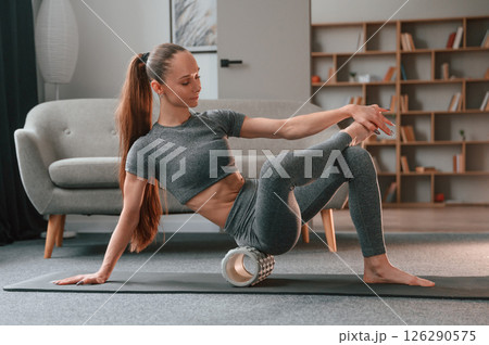 Sitting on the foam roller and smiling. Young woman in yoga clothes doing fitness indoors Sitting on the foam roller and smiling. Young woman in yoga clothes doing fitness indoors 126290575