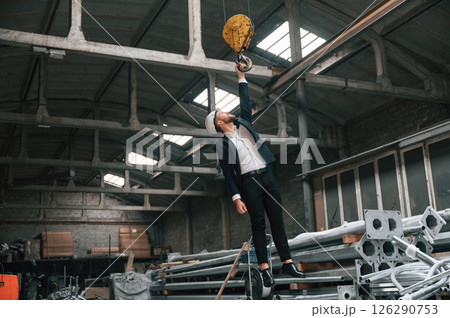 Having fun by hanging on the crane's hook. Manager in black formal wear at the factory 126290753