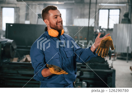 In yellow colored hear protective headphones and gloves. Factory worker in blue uniform is indoors 126290780