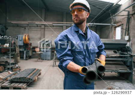 Looking to the side and holding metal pipes. Factory worker in blue uniform is indoors 126290937