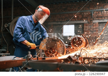 In protective shield on head. Welding the metal. Factory worker in blue uniform is indoors 126290947
