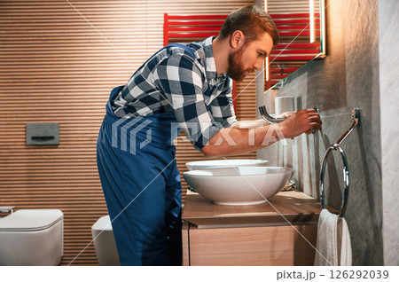 Uses a screwdriver for repairs. Plumber in blue uniform is at work in the bathroom Uses a screwdriver for repairs. Plumber in blue uniform is at work in the bathroom 126292039