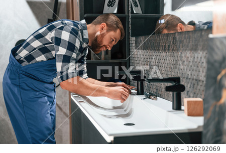 Side view. Fixing water tap. Plumber in blue uniform is at work in the bathroom Side view. Fixing water tap. Plumber in blue uniform is at work in the bathroom 126292069