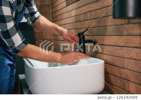 Standing by sink and fixing water tap. Plumber in blue uniform is at work in the bathroom Standing by sink and fixing water tap. Plumber in blue uniform is at work in the bathroom 126292099