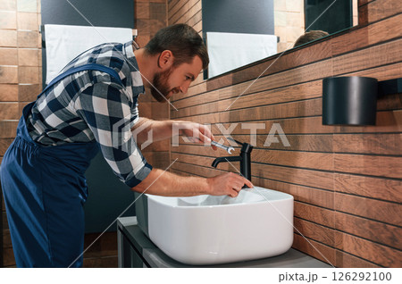 Standing by sink and fixing water tap. Plumber in blue uniform is at work in the bathroom 126292100