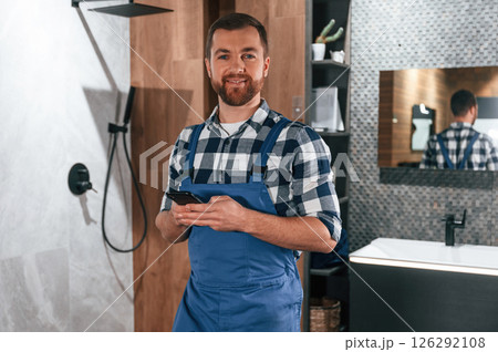 Standing and holding smartphone. Plumber in blue uniform is at work in the bathroom 126292108