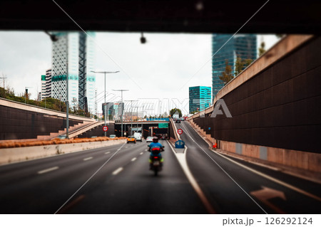 Vehicles drive through a tunnel on a multi-lane highway, buildings lining the sides, cloudy skies overhead. The perspective is from the driver's seat. 126292124