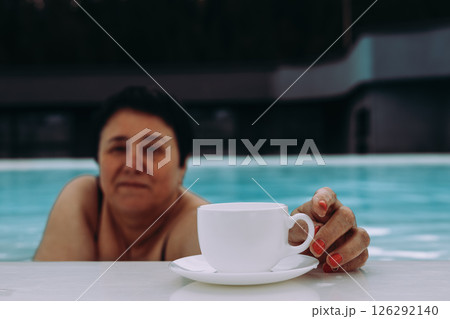 A relaxed woman enjoying a cup of coffee while relaxing in a refreshing swimming pool. A woman in a pool with a cup of tea. Relaxing moment in water. 126292140
