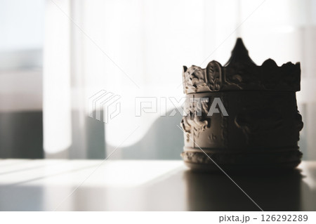 A crown-shaped trinket box sits on a reflective surface, partially illuminated by sunlight filtering through nearby window. Porcelain carousel indoors 126292289