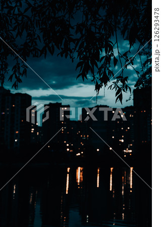 A serene cityscape at dusk, where the illuminated windows of buildings reflect beautifully on the water. Overhanging leaves frame the sky above. 126293478