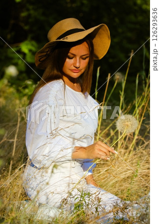 A beautiful gorgeous young woman in a white dress and sun hat poses among tall grass, holding a dandelion, bathed in sunlight. Summertime vacation. A beautiful gorgeous young woman in a white dress and sun hat poses among tall grass, holding a dandelion, bathed in sunlight. Summertime vacation. 126293636