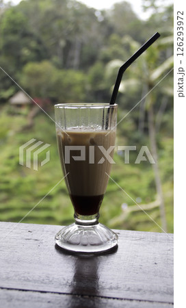 Iced latte in a tall glass with a black straw on a wooden table with blurred tropical greenery and rice terrace view in Bali Iced latte in a tall glass with a black straw on a wooden table with blurred tropical greenery and rice terrace view in Bali 126293972