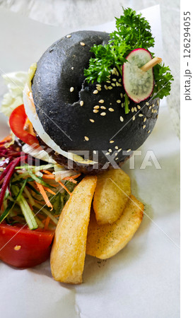 Gourmet black charcoal burger with egg, cheese, and vegetables, served with crispy wedges and fresh salad on parchment, creating a vibrant and appetizing presentation. 126294055