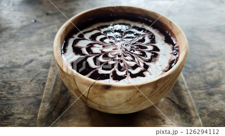 Elegant bowl of black glutinous rice porridge drizzled with intricate coconut milk art, served in a rustic wooden bowl on a textured wooden surface for a traditional Indonesian dessert experience. Elegant bowl of black glutinous rice porridge drizzled with intricate coconut milk art, served in a rustic wooden bowl on a textured wooden surface for a traditional Indonesian dessert experience. 126294112