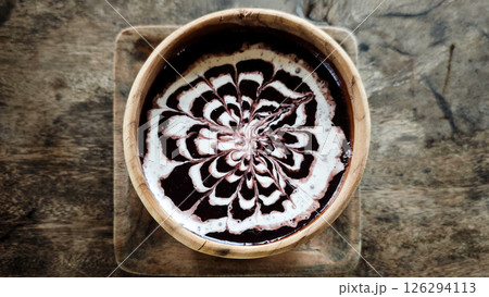 Elegant bowl of black glutinous rice porridge drizzled with intricate coconut milk art, served in a rustic wooden bowl on a textured wooden surface for a traditional Indonesian dessert experience. 126294113
