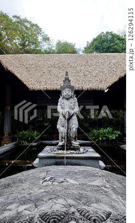Traditional Balinese Water Fountain Statue with Stone Carvings and Thatch Roof Pavilion Surrounded by Greenery in Tropical Garden Setting 126294115