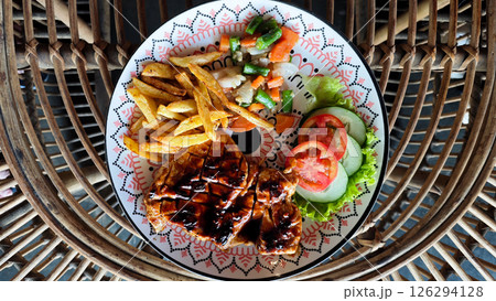 Grilled Chicken Steak with French Fries and Fresh Vegetable Salad on a Decorative Plate with Wicker Furniture Background Grilled Chicken Steak with French Fries and Fresh Vegetable Salad on a Decorative Plate with Wicker Furniture Background 126294128