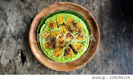 Vibrant green banana pancake with caramelized banana slices and brown sugar, served on a wooden plate with a rustic dark wood background 126294134