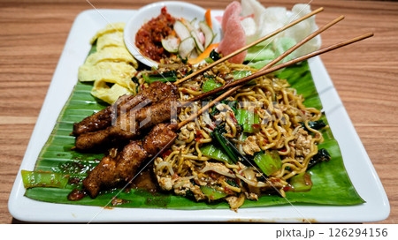 Stir-fried noodles served with grilled chicken skewers, omelette slices, and assorted vegetables on a banana leaf-lined plate with sambal and crackers on the side, presented on a wooden table 126294156