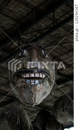 Old Coconut Carved into a Scary Face Hanging from the Roof in a Dimly Lit Area with Bamboo Ceiling Background 126294167