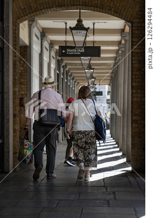 Vertical view of An elderly couple holding hands walking through the symmetrical arches and old fashioned street lamps hang from the ceiling at Covent Garden in London. Vertical view of An elderly couple holding hands walking through the symmetrical arches and old fashioned street lamps hang from the ceiling at Covent Garden in London. 126294484