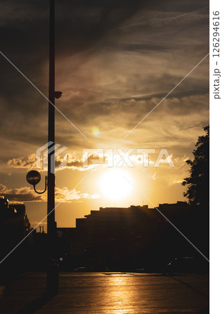 Buildings silhouetted against a sunset, a street lamp stands tall, casting shadows across the urban landscape. The sun provides a beautiful warm glow. 126294616