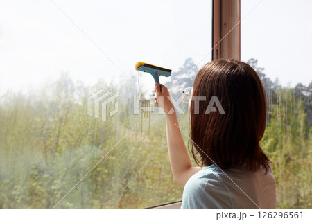 A young women is cleaning a large window with a squeegee, revealing a scenic outdoor view. A young women is cleaning a large window with a squeegee, revealing a scenic outdoor view. 126296561