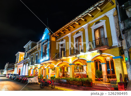 A historic colonial-era building with arched porticoes and balconies glows under streetlights at the Central Park of Granada, Nicaragua. The nighttime setting highlights the rich yellow facade and 126298495