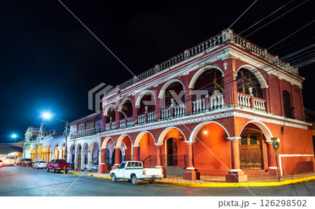 A historic colonial-era building with arched porticoes and balconies glows under streetlights in the heart of Granada, Nicaragua. The nighttime setting highlights the rich red facade and architectural 126298502