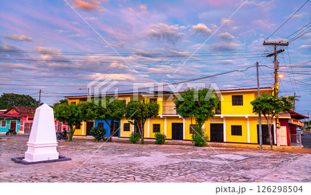 Small cobblestone plaza with neatly pruned trees, a white obelisk, and brightly painted yellow buildings, set near the historic Fortaleza de la Polvora in Granada, Nicaragua, under a vivid pink and 126298504