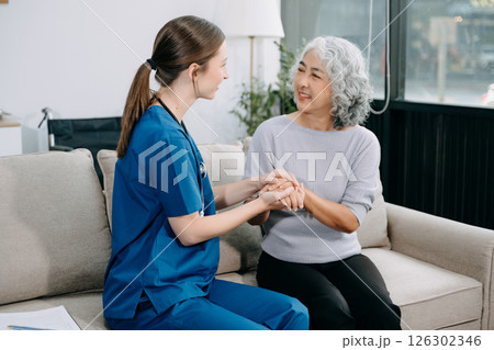 Young nurse helping elderly woman walk in the room, holding his hand, supporting. Treatment and rehabilitation after injury in assisted living facility 126302346