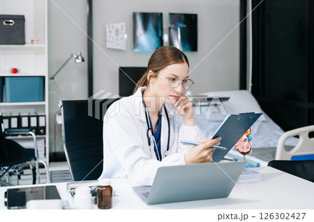 Confident young Caucasian female doctor in white medical uniform sit at desk working on computer. Smiling use laptop write in medical journal in clinic. 126302427