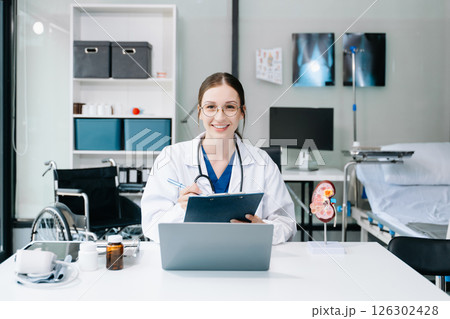 Young female doctor in white medical uniform using laptop and tablet talking video conference call at desk,Doctor sitting at desk and writing a prescription for her patient Young female doctor in white medical uniform using laptop and tablet talking video conference call at desk,Doctor sitting at desk and writing a prescription for her patient 126302428
