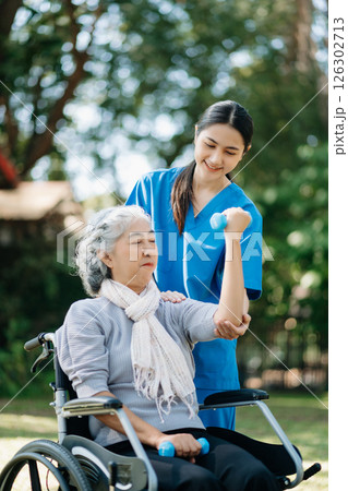 Asian physiotherapist helping elderly woman patient stretching arm during exercise correct with dumbbell in hand during training hand with patient Back problems in the garden. 126302713