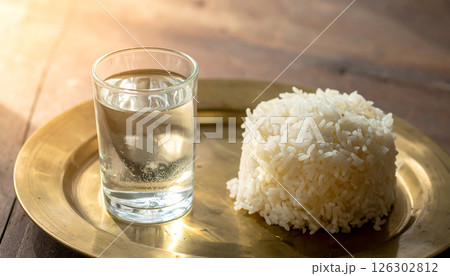 A glass of water and a mound of white rice on a brass plate, illuminated by warm light. 126302812
