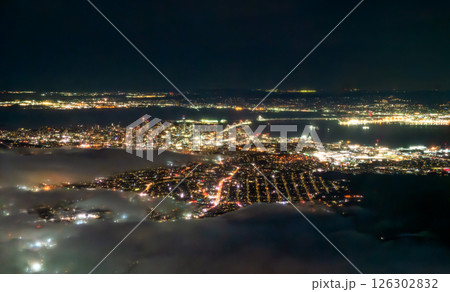 Aerial night view of San Francisco glowing with city lights under a partially foggy sky, with the Bay Bridge illuminated across the bay and the city grid extending into the distance. The shimmering 126302832