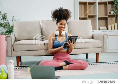 Active woman in athleisure sitting on yoga mat with tablet, headphones and modern home lifestyle. Active woman in athleisure sitting on yoga mat with tablet, headphones and modern home lifestyle. 126303489