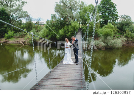 Beautiful newlywed couple in love posing on suspension bridge against summer nature background Beautiful newlywed couple in love posing on suspension bridge against summer nature background 126303957