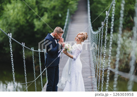 Beautiful newlywed couple in love posing on suspension bridge against summer nature background Beautiful newlywed couple in love posing on suspension bridge against summer nature background 126303958