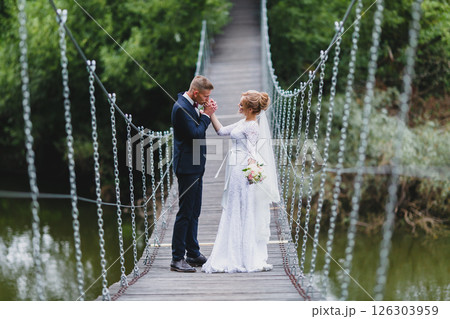 Beautiful newlywed couple in love posing on suspension bridge against summer nature background Beautiful newlywed couple in love posing on suspension bridge against summer nature background 126303959