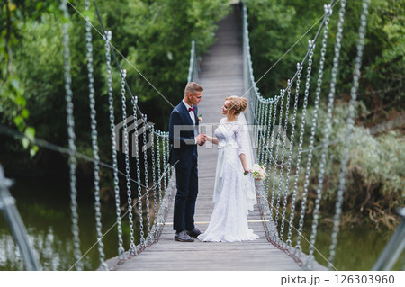 Beautiful newlywed couple in love posing on suspension bridge against summer nature background Beautiful newlywed couple in love posing on suspension bridge against summer nature background 126303960