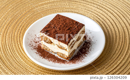 A slice of classic Tiramisu dessert on a white plate, dusted with cocoa, sits on a golden textured placemat. A slice of classic Tiramisu dessert on a white plate, dusted with cocoa, sits on a golden textured placemat. 126304209