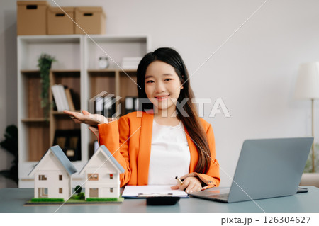 Young real estate agent worker working with laptop and tablet at table in office and small house beside it. Young real estate agent worker working with laptop and tablet at table in office and small house beside it. 126304627