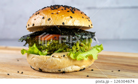 A vegetarian burger with a green patty, seaweed, lettuce, and tomato on a bun with black sesame seeds, placed on a wooden board. 126304674