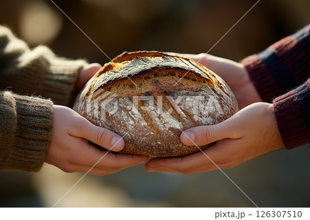 Close-up of four hands holding a rustic sourdough loaf outdoors, crust cracked and golden brown 126307510