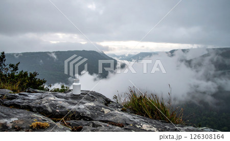 A white bottle sits on a rock overlooking a misty mountain valley under a cloudy sky, creating a serene and atmospheric scene. A white bottle sits on a rock overlooking a misty mountain valley under a cloudy sky, creating a serene and atmospheric scene. 126308164