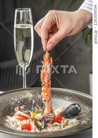 Hand holding a grilled shrimp over a gourmet seafood risotto with a glass of champagne. Hand holding a grilled shrimp over a gourmet seafood risotto with a glass of champagne. 126309594