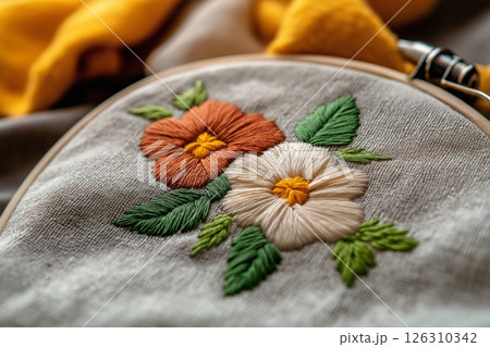 An embroidery hoop with two detailed flowers in earthy tones, surrounded by green stitched leaves on fabric. 126310342
