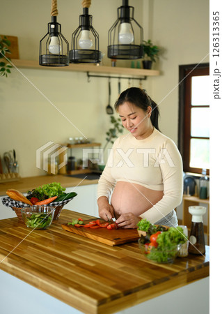 Happy pregnant woman stands at kitchen counter, preparing a wholesome meal Happy pregnant woman stands at kitchen counter, preparing a wholesome meal 126313365